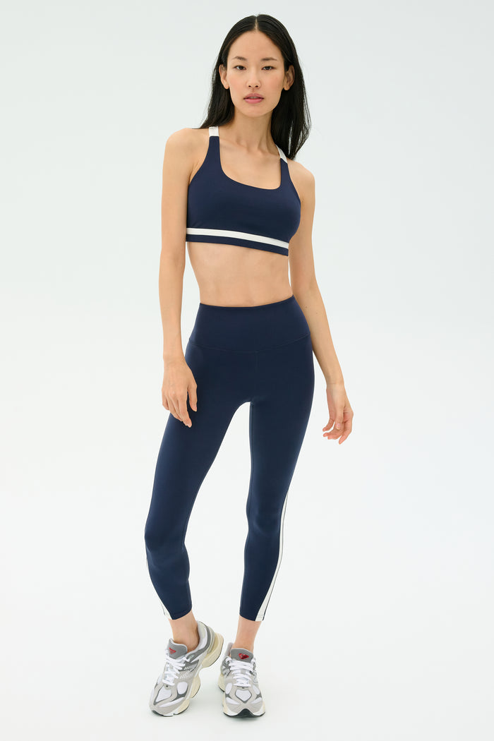 A woman faces forward wearing the SPLITS59 Miles Rigor Bra in Indigo/White, styled with matching leggings and silver sneakers.