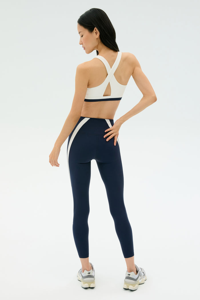 A woman faces away, wearing the SPLITS59 Miles Rigor Bra in Indigo/White, paired with leggings and white sneakers against a plain light background.