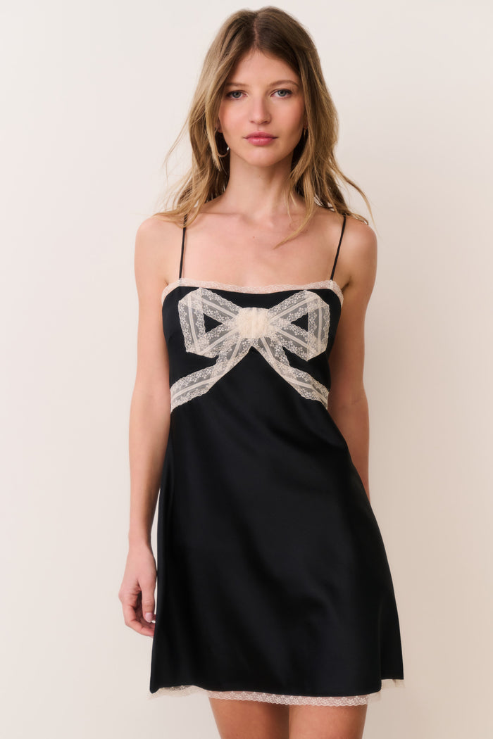 Woman wearing a black satin dress with white lace trim and bow detail against a plain background