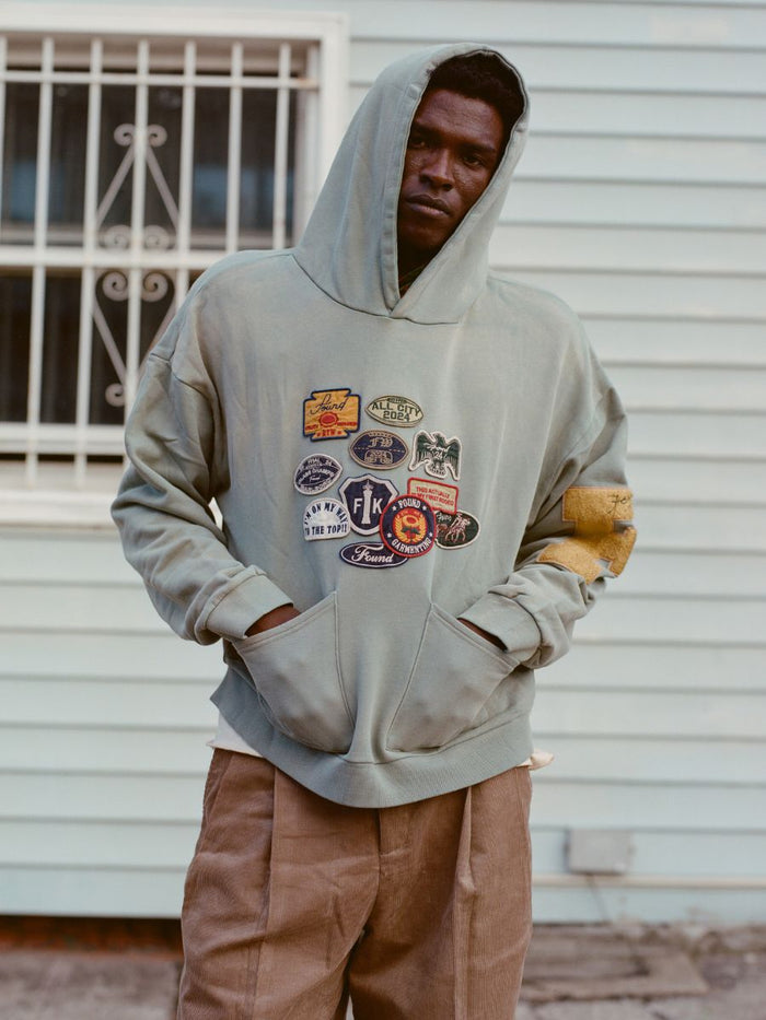 In front of a light blue building with a barred window, someone stands wearing FOUND's Multi Patch Hoodie, adorned with embroidered patches.