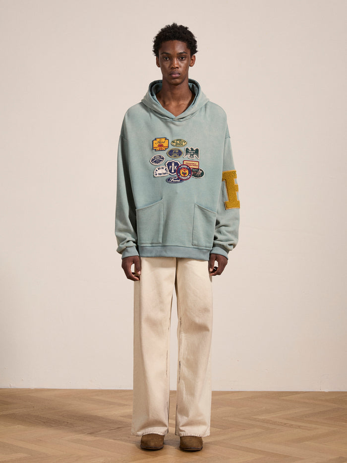 A person standing indoors, wearing FOUND's Multi Patch Hoodie crafted from vintage-inspired French Terry cotton in sage green, adorned with multiple patches on the front and a large letter "E" on the sleeve, paired with off-white pants and brown shoes.