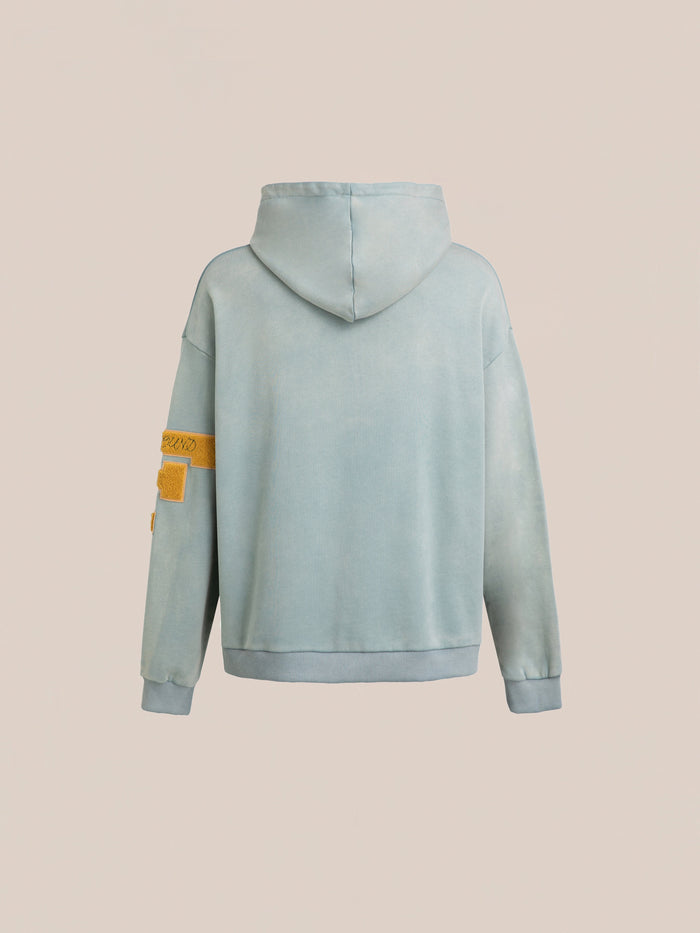 A back view of the Multi Patch Hoodie by FOUND in light blue, crafted from soft French Terry cotton, featuring a hood and a vintage-inspired orange patch on the left sleeve.