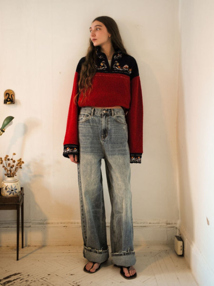 Indoors, an individual models a red and black FOUND Pahari Floral Embroidered Sherpa Pullover paired with baggy jeans and sandals. In the background, a small table holds florally embroidered plants.