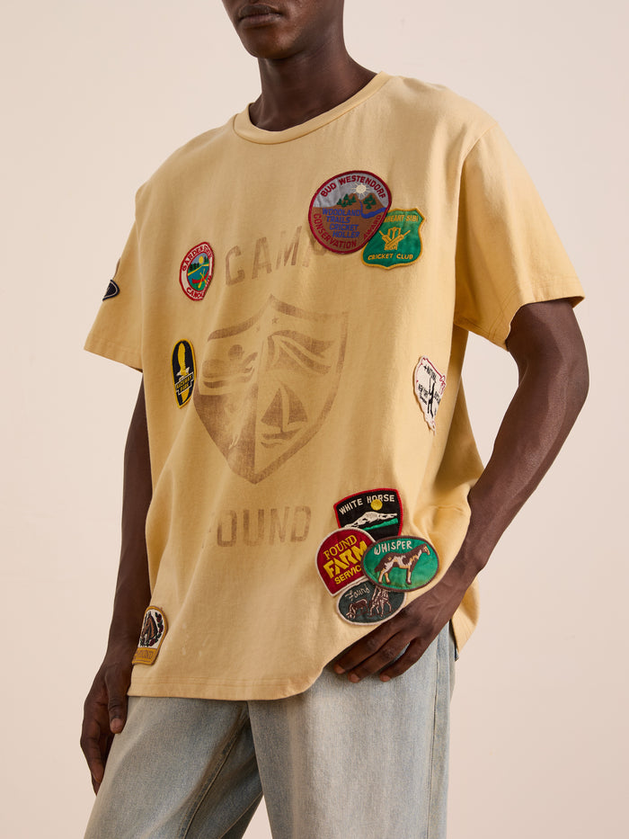 Someone in a FOUND Patchwork Camp Tee—unisex, relaxed fit, yellow with vintage-style embroidered patches—paired with light blue jeans.