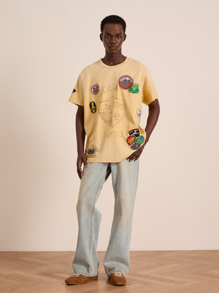 Indoors, a person wears the FOUND Patchwork Camp Tee in pale yellow with vintage-inspired embroidered patches, paired with light blue jeans and brown shoes, standing against a plain background.
