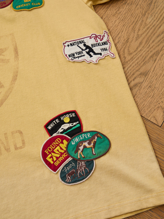 The FOUND Patchwork Camp Tee is a unisex relaxed-fit yellow tee featuring vintage-inspired embroidered patches of maps, animals, and club logos, shown on a wooden floor.