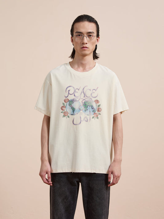 A person with long dark hair and glasses wears the FOUND Peace Globe Tee, a cream vintage cotton t-shirt with floral graphics and a unisex relaxed fit, standing against a plain beige background.