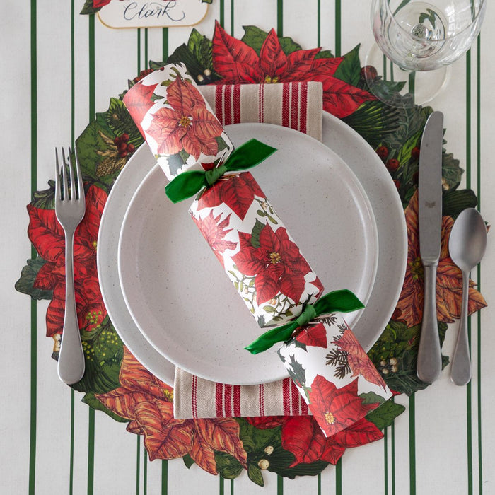 Christmas-themed table setting with poinsettia napkin, greenery placemat, and cutlery on a striped background.