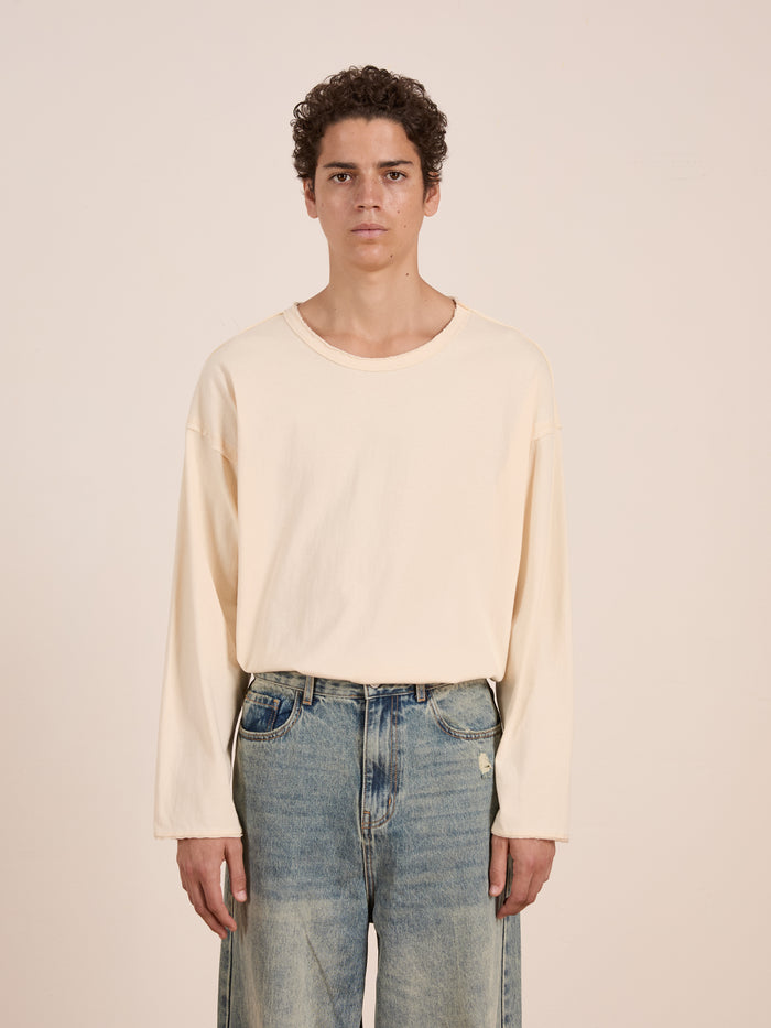 A person with short curly hair wears the FOUND Reversed LS Tee—loose-fitting with a distressed neckline—and light blue jeans, standing against a plain off-white background.
