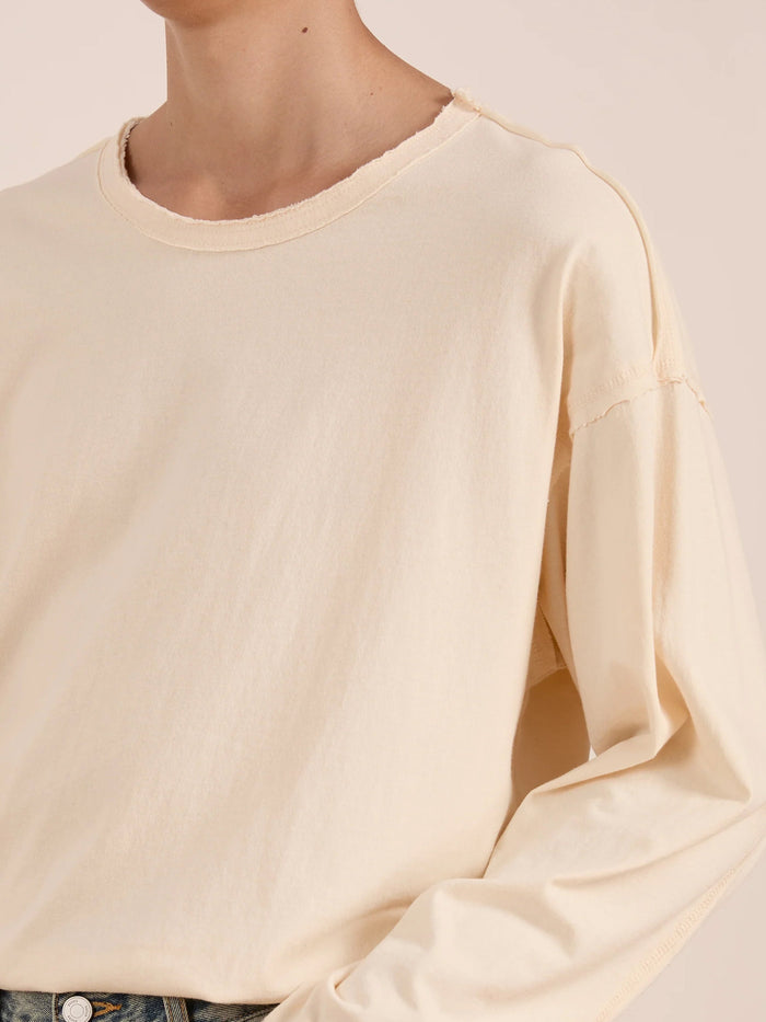 A person wearing the FOUND Reversed LS Tee, a unisex off-white long sleeve top featuring exposed seams and a distressed neckline, shown from shoulders to waist.