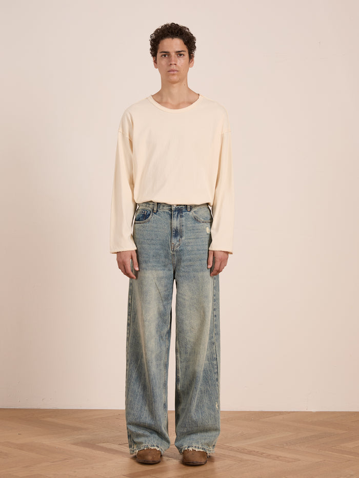 Indoors against a plain wall, a person wears FOUND’s Reversed LS Tee—a loose unisex long sleeve with a distressed neckline—paired with wide-leg faded jeans and brown shoes.