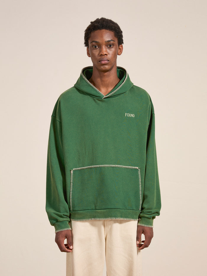 A person faces forward wearing the FOUND Stitched Border Distressed Logo Hoodie in green French terry cotton with light stitching, paired with light pants, against a neutral background.
