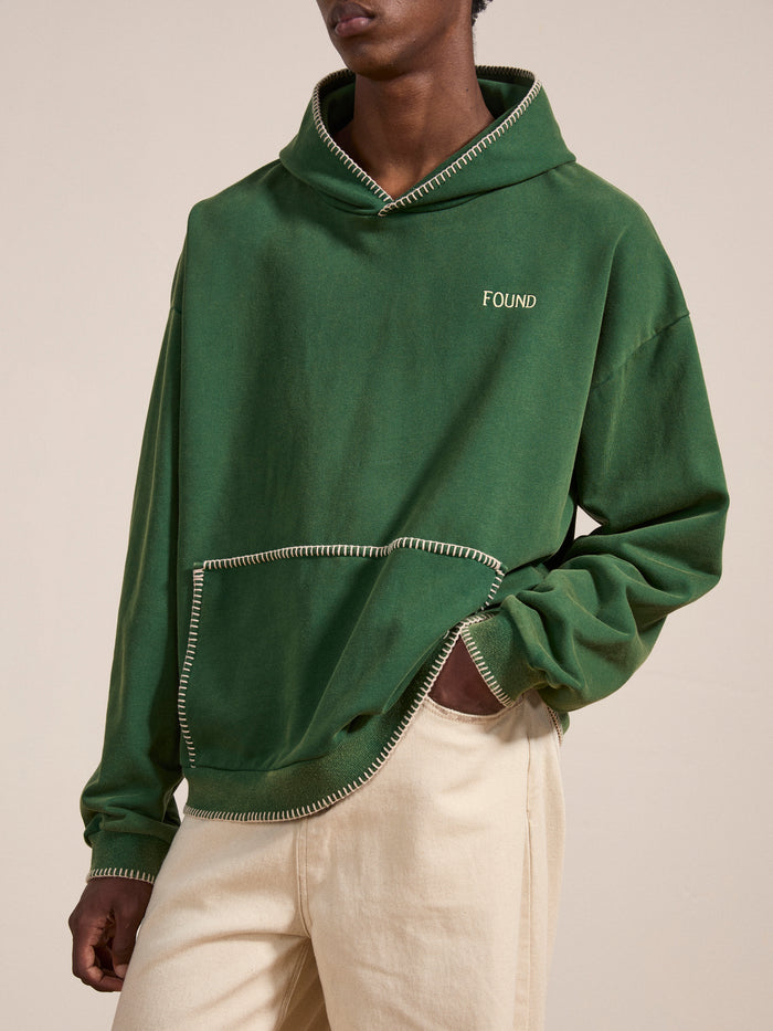 Someone wearing the FOUND Stitched Border Distressed Logo Hoodie—an oversized green French terry cotton piece with white stitching and an embroidered logo on the chest—paired with beige pants.
