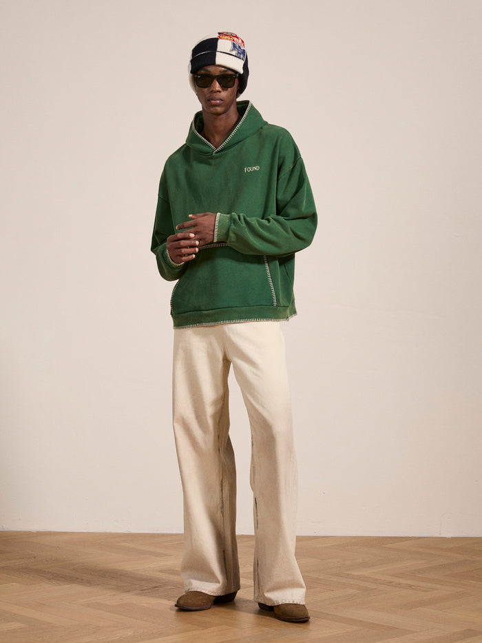 A person wears FOUND's Stitched Border Distressed Logo Hoodie in green, paired with white wide-leg pants, brown shoes, sunglasses, and a headscarf, standing indoors against a plain background.