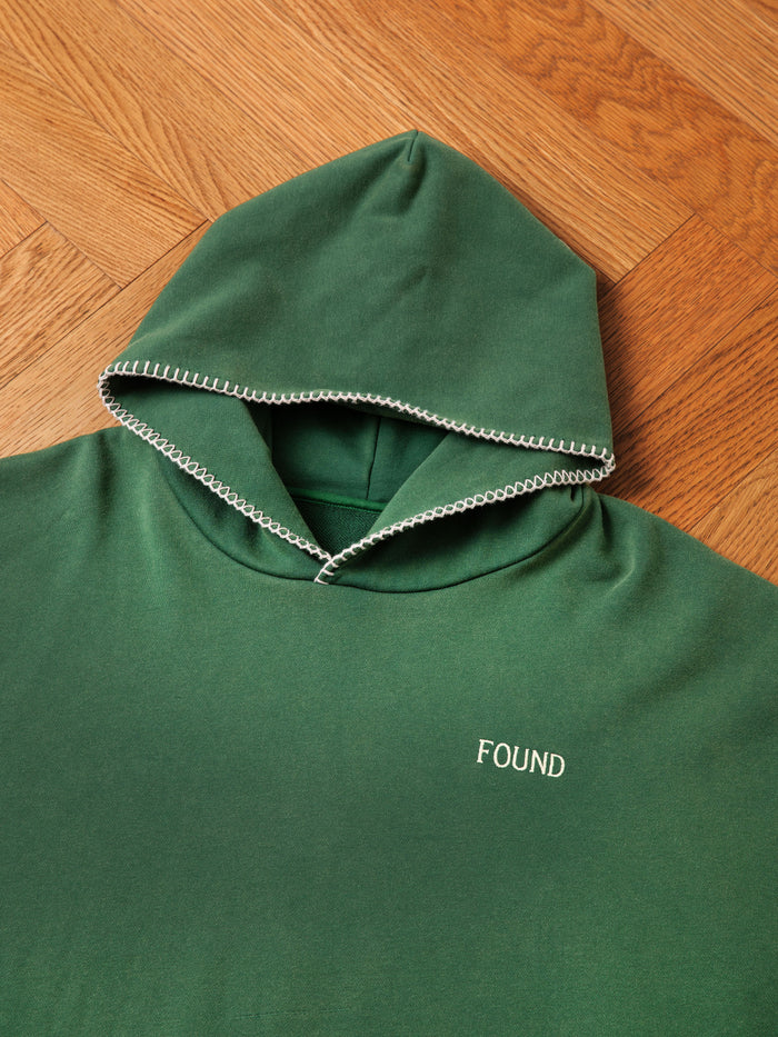 The Stitched Border Distressed Logo Hoodie by FOUND, made from soft French terry cotton, features white hood-edge stitching and "FOUND" embroidered on the chest. Displayed laid flat on a wooden floor.