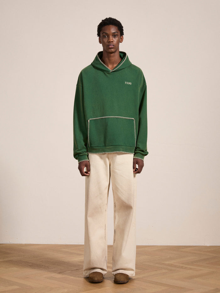 A person stands indoors against a plain background wearing the FOUND Stitched Border Distressed Logo Hoodie in vintage oversized green, paired with cream wide-leg pants and brown shoes.