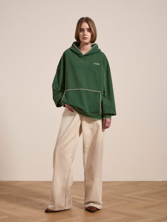 A woman stands indoors on a wooden floor wearing the FOUND Stitched Border Distressed Logo Hoodie in vintage green and wide beige pants, set against a plain light-colored wall.