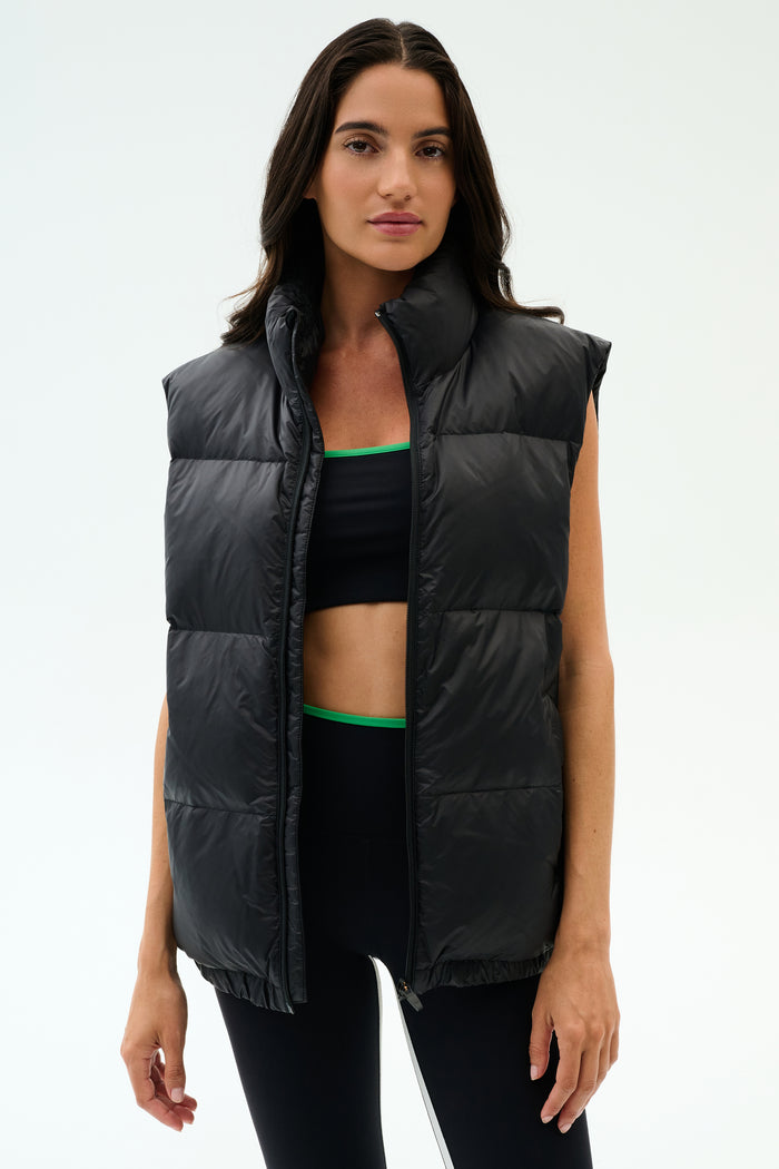 A woman wears the SPLITS59 Summit Nylon Puffer Vest in black over a sports bra and leggings, standing against a plain light background—the perfect lightweight layer for cool weather workouts.