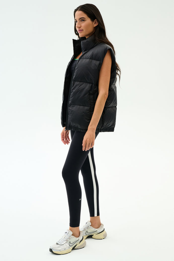 A woman stands in profile against a white background wearing the SPLITS59 Summit Nylon Puffer Vest in black, paired with black leggings featuring a white stripe and white athletic sneakers—highlighting its lightweight, cool-weather insulation for workouts.