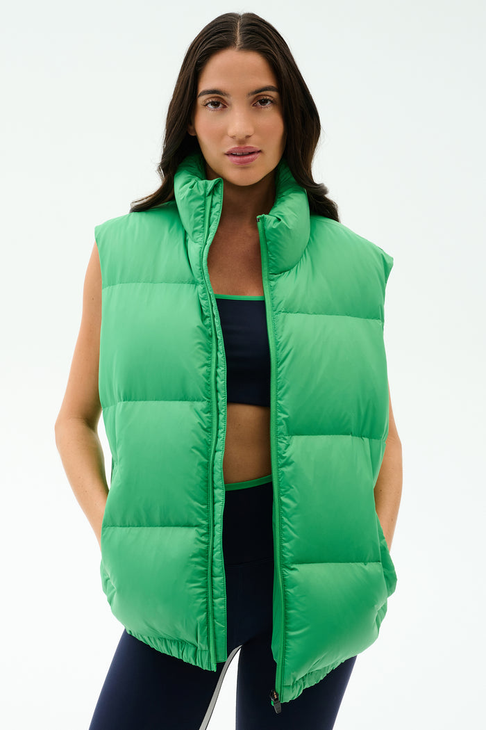 Wearing the SPLITS59 Summit Nylon Puffer Vest in Grass over a dark sports bra and leggings, this person stands against a plain light background—ready for cool weather workouts with lightweight warmth and style.
