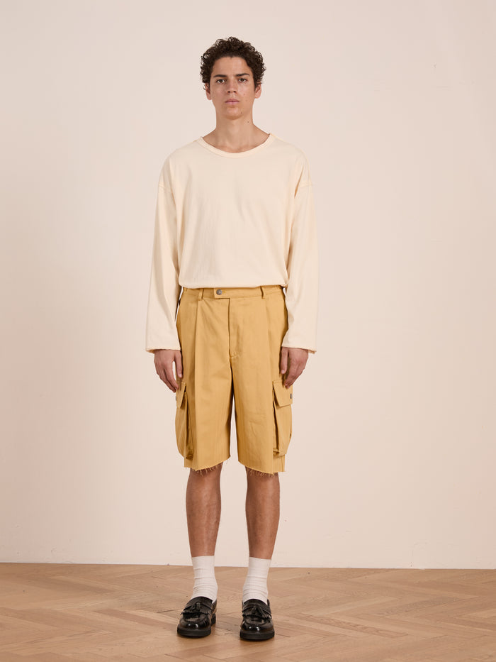 A young man stands indoors against a plain background in relaxed, unisex style, wearing FOUND Sunlight Twill Cargo Shorts with a cream herringbone shirt, white socks, and black loafers.