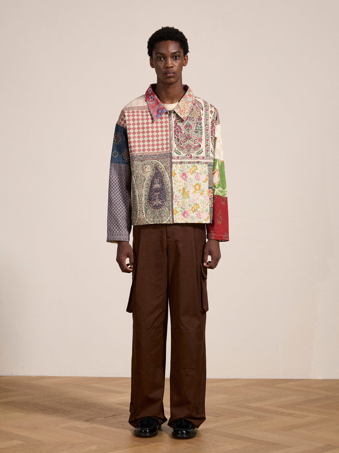 A person stands against a plain wall wearing the FOUND Tapestry Twill Jacket adorned with South Asian patterns and brown cargo pants, looking at the camera.