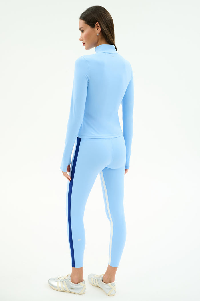 A woman wearing the SPLITS59 Tess Rigor Half Zip Waist Length jacket in Skylight with matching leggings and white sneakers, standing and facing away on a plain background.