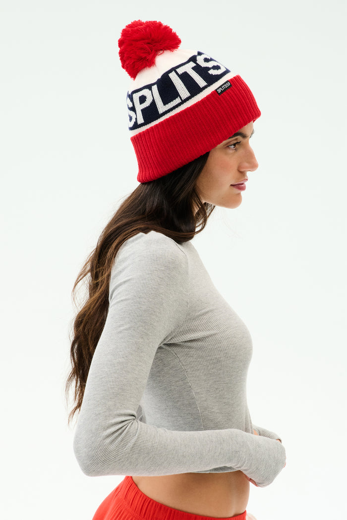 Woman in profile wearing a SPLITS59 Winter Pom Pom Beanie - Lotus Red/White with "SPLITS" text, a gray long-sleeve top, and red pants—perfect for winter workouts against a white background.