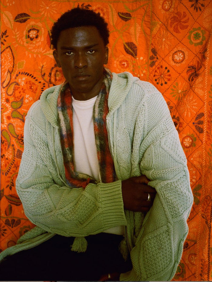 A man sits against an orange patterned backdrop, wrapped in a mint green Zip Up Cable Knit Hoodie by FOUND, layered over a white shirt and accented with a multicolored plaid scarf.