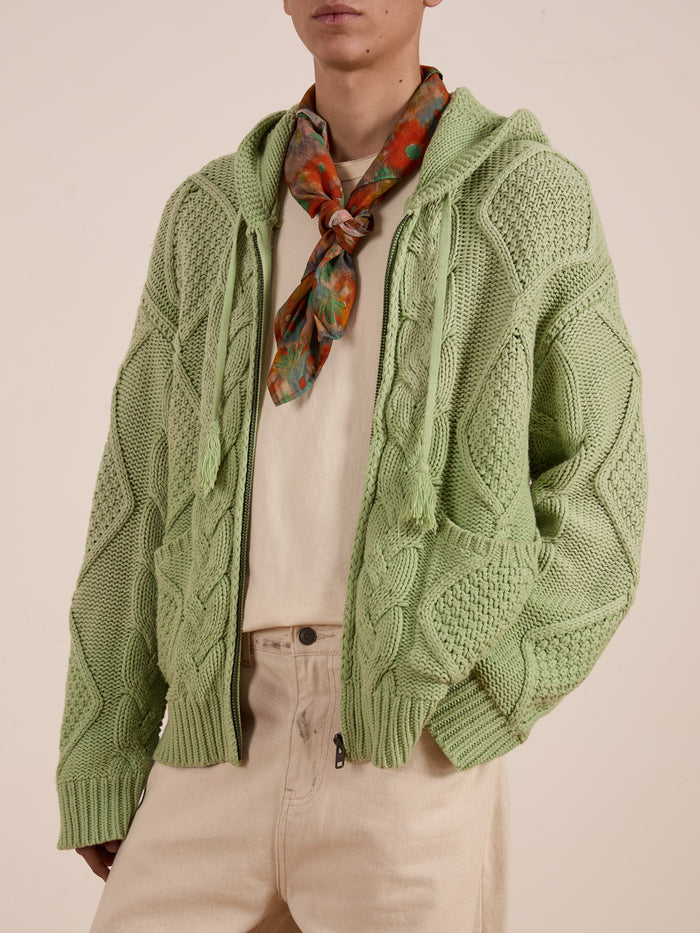 A person is wearing a green FOUND Zip Up Cable Knit Hoodie in a unisex relaxed fit, paired with a beige shirt, beige pants, and a multicolored neck scarf.